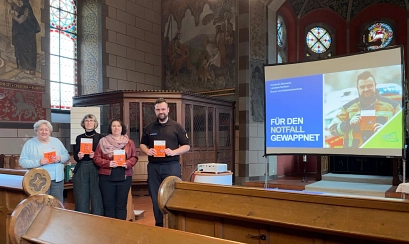 Zu sehen sind vier Personen, drei Frauen und ein Mann, die in einer Kirche stehen. Im Vordergund sieht man Kirchenbänke. Alle vier halten eine organgefarbene Broschüre in den Händen und schauen in die Kamera&nbsp;&copy;&nbsp;Landkreis Northeim