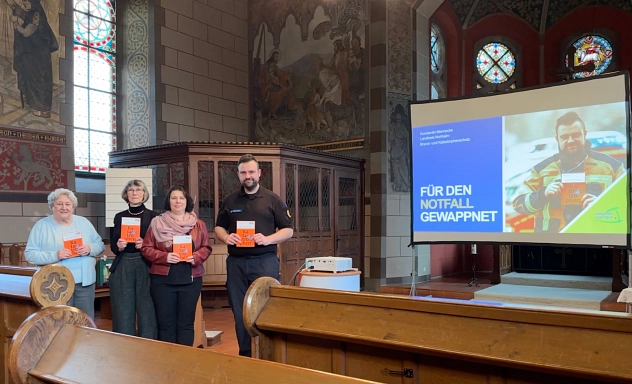 1. Veranstaltung "Älter werden und dabei das Gute für sich bewahren" Zu sehen sind vier Personen, drei Frauen und ein Mann, die in einer Kirche stehen. Im Vordergund sieht man Kirchenbänke. Alle vier halten eine organgefarbene Broschüre in den Händen und schauen in die Kamera