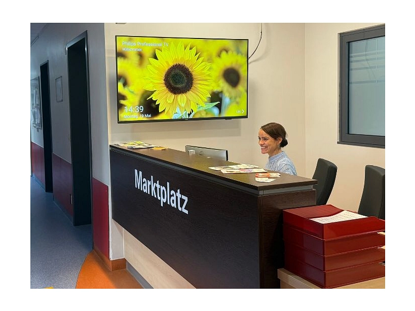 Zu sehen ist die Anmeldung der psychiatrischen Vesorgungseinheit der UMG im Bürgergesundheitspark Bad Gandersheim. Auf dem Tresen steht "Marktplatz", dahinter sitzt eine junge Frau. Im Hintergrund ist ein Bildschirm zu sehen, auf dem gerade ein Bild von einer Sonnenblume projiziert ist,