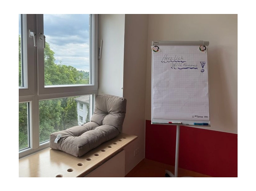 Zu sehen ist eine breite Fensterbank, darauf liegt ein Kissen, auf das man sich setzen kann. Daneben steht ein Flipchart auf das "Herzlich Willkommen" geschrieben steht.