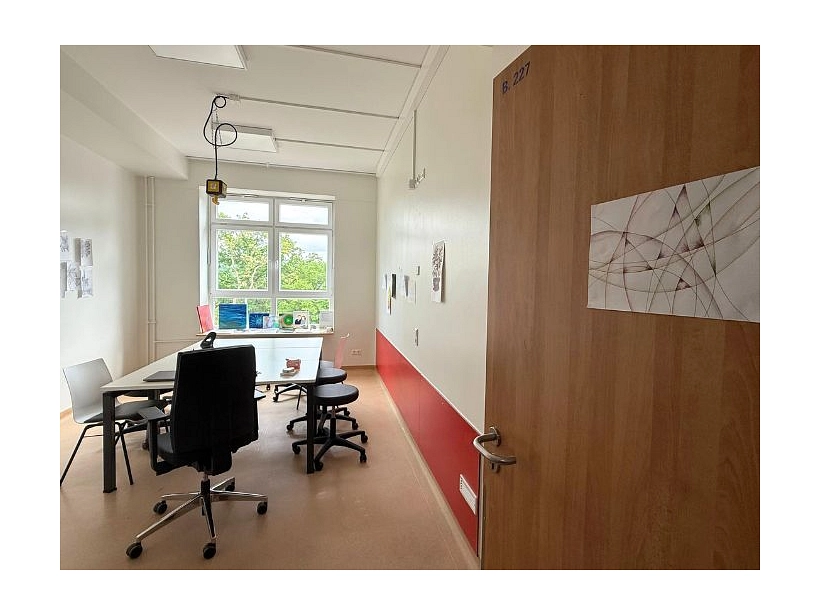 Zu sehen ist eine offene Tür an der ein gezeichnetes abstraktes Bild hängt. Hinter der Tür ist ein Kunstraum mit einem großen Tisch und mehreren Stühlen. Es hängen selbstgemalte Bilder an der Wand.