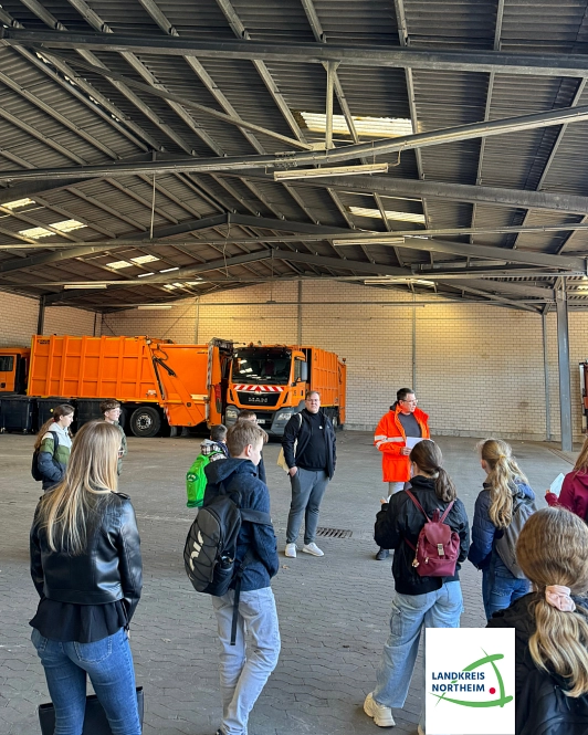 Zu sehen sind einige Jungen und Mädchen sowie ein Mitarbeiter der Kreisabfallwirtschaft, alle stehen in einer großen Halle, in der im Hintergrund auch ein Müllfahrzeug zu sehen ist.