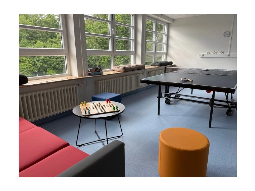 Zu sehen ist ein Zimmer mit einer Tischtennisplatte, einen Sofa, einem Tisch mit Mensch ärgere Dich nicht Spiel und mehreren Hockern.