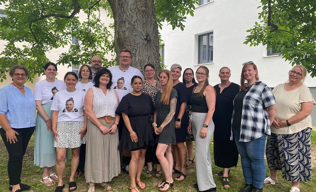 Absolvent*innen Schulbegleitung KVHS Eine Gruppe von 16 Personen steht im Freien vor einem großen Baum und einem hellen Gebäude.