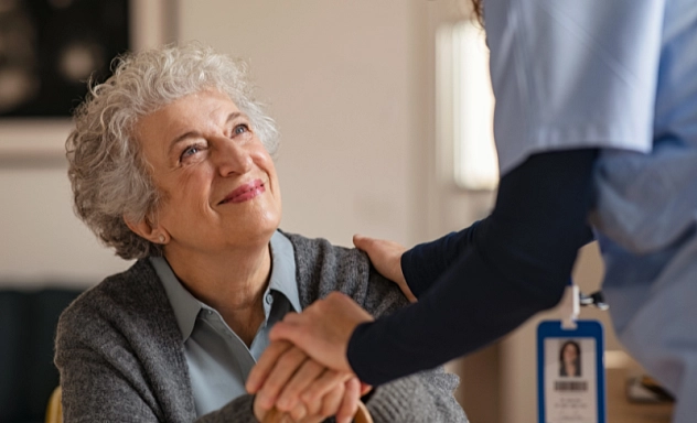 Zu sehen ist eine Rentnerin die sitzt, und die mit einer Pflegeperson spricht, die eine Hand auf die Schulter der Rentnerin legt.