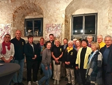 Auftaktveranstaltung "Die Germanen - ein Mythos?" Das Foto zeigt eine Gruppe von etwa 15 Personen, die in einem historischen Gewölberaum mit Steinwänden und hohen Fenstern zusammenstehen. Einige tragen dunkle Polohemden mit einem gelben Emblem, andere Freizeitkleidung in helleren Farben. Im Hintergrund sind Kunstwerke mit floralen Motiven an den Wänden zu sehen.