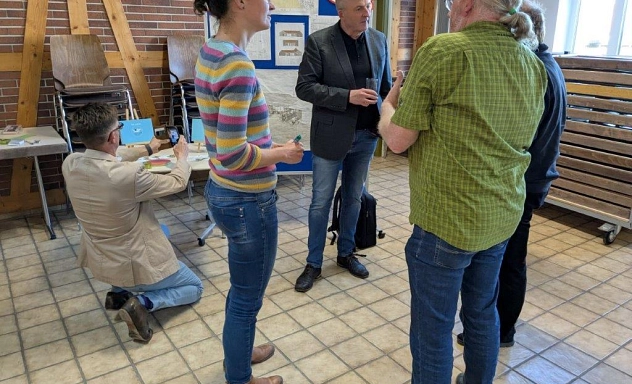 Austauschveranstaltung bürgerschaftlichen Initiativen in Hevensen 05.2025 Mehrere Personen unterhalten sich in einem Raum mit gefliesten Boden und Backsteinwänden. Eine Frau im Vordergrund trägt einen bunten, gestreiften Pullover, ein Mann in einem grünen Hemd spricht mit ihr. Ein Mann im grauen Sakko hält ein Glas und hört zu. Im Hintergrund kniet ein Mann mit hellem Sakko und macht ein Foto von einem Tisch mit Flyern. Stühle sind an der Wand gestapelt, und eine Pinnwand mit Plakaten steht hinten.