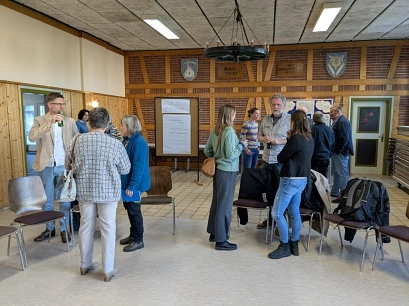 Mehrere Menschen stehen in kleinen Gruppen in einem Gemeinschaftsraum mit Holz- und Backsteinwänden und unterhalten sich. Einige trinken etwas, andere hören zu oder sprechen miteinander. Im Hintergrund hängen Wappen an der Wand, und eine Flipchart mit handgeschriebenem Text steht vorne. Auf der rechten Seite sind Stühle aufgestellt, einige mit Jacken und Taschen belegt.