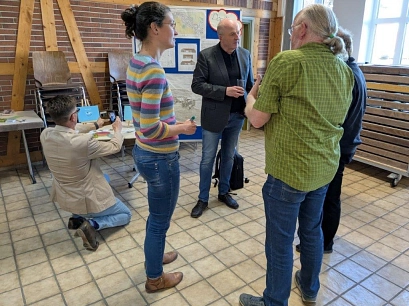 Mehrere Personen unterhalten sich in einem Raum mit gefliesten Boden und Backsteinwänden. Eine Frau im Vordergrund trägt einen bunten, gestreiften Pullover, ein Mann in einem grünen Hemd spricht mit ihr. Ein Mann im grauen Sakko hält ein Glas und hört zu. Im Hintergrund kniet ein Mann mit hellem Sakko und macht ein Foto von einem Tisch mit Flyern. Stühle sind an der Wand gestapelt, und eine Pinnwand mit Plakaten steht hinten.