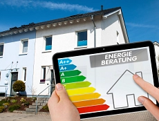 Im Vordergrund ist ein Tablet zu sehen, auf dem die Skizze eines Hauses und die Energieklassen zu sehen sind. Darüber steht "Energieberatung". Im Hintergrund des Bildes sind zwei Reihenhäuser zu sehen.
