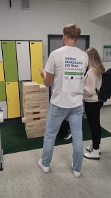 Zwei Personen stehen vor einem übergroßen Jenga-Turm aus Holzklötzen in einem Schulgebäude. Eine Person trägt ein T-Shirt mit der Aufschrift ‚Mobiles Demokratiezentrum im Landkreis Northeim – Demokratie vor Ort!‘. Auf den Holzklötzen stehen Begriffe wie ‚Partizipation‘. Das Spiel dient als Mitmachaktion, um mit Besucherinnen und Besuchern über demokratische Werte und gesellschaftlichen Zusammenhalt ins Gespräch zu kommen.&nbsp;&copy;&nbsp;Landkreis Northeim