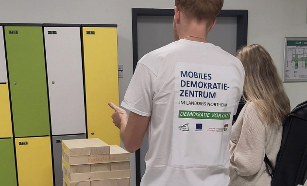 Berufsinformationsmarkt2026_Mobiles Demokratiezentrum_quer.jpg Zwei Personen stehen vor einem übergroßen Jenga-Turm aus Holzklötzen in einem Schulgebäude. Eine Person trägt ein T-Shirt mit der Aufschrift ‚Mobiles Demokratiezentrum im Landkreis Northeim – Demokratie vor Ort!‘. Auf den Holzklötzen stehen Begriffe wie ‚Partizipation‘. Das Spiel dient als Mitmachaktion, um mit Besucherinnen und Besuchern über demokratische Werte und gesellschaftlichen Zusammenhalt ins Gespräch zu kommen.