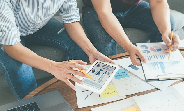 Das Bild zeigt zwei Personen, die an einem Tisch sitzen und über eine Auswahl an Geschäftsdokumenten und Diagrammen diskutieren. Eine Person hält ein Tablet, auf dem ein Diagramm angezeigt wird, während die andere Person auf ein Papier mit einem Stift zeigt. Auf dem Tisch liegen verschiedene Dokumente mit Diagrammen, Notizen und einem Laptop, die auf eine intensive Besprechung oder Planungsarbeit hindeuten.