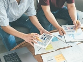 Das Bild zeigt zwei Personen, die an einem Tisch sitzen und über eine Auswahl an Geschäftsdokumenten und Diagrammen diskutieren. Eine Person hält ein Tablet, auf dem ein Diagramm angezeigt wird, während die andere Person auf ein Papier mit einem Stift zeigt. Auf dem Tisch liegen verschiedene Dokumente mit Diagrammen, Notizen und einem Laptop, die auf eine intensive Besprechung oder Planungsarbeit hindeuten.&nbsp;&copy;&nbsp;canva