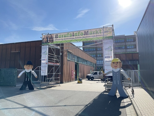 Zu sehen ist der Eingang mit dem Banner "Berufsinfomarkt". Links und rechts davon steht jeweils eine große Figur in Handwerkerkleidung.