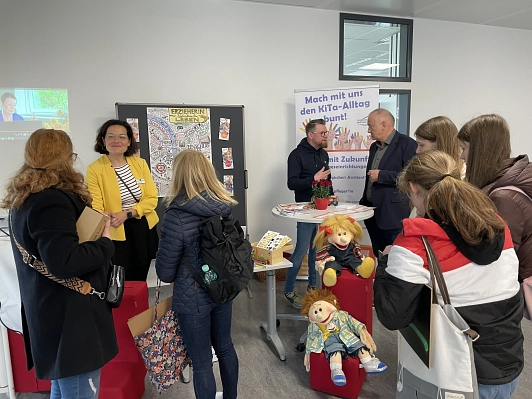 Vor dem Stand des Landkreises Northeim zum Thema "Mach mit uns den KiTa-Alltag bunt" stehen einige Schülerinnen, um sich zu informieren. Vor dem Stand sind außerdem zwei große Stoffpuppen drapiert.
