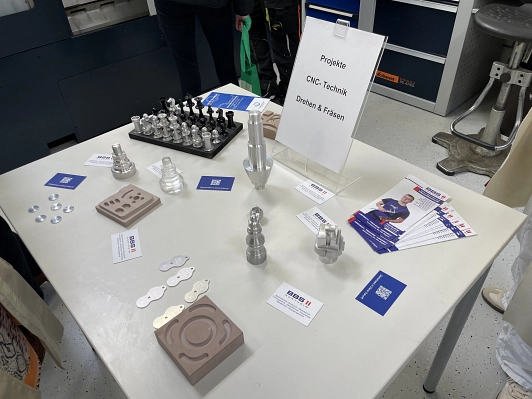 Auf einem Tisch sind Gegenstände ausgestellt, die in der Abteilung "Drehen und Fräsen" entstanden sind, zum Beispiel Einkaufschips aus Metall oder ein Schachset.