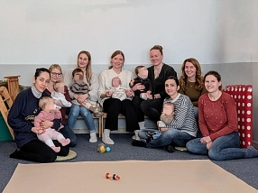 Eine Gruppe von acht Frauen sitzt gemeinsam mit ihren Babys in einem freundlichen Raum mit Teppichboden. Einige Mütter halten ihre Kinder auf dem Schoß, während andere auf einer Bank oder dem Boden sitzen. Die Babys tragen bequeme Kleidung und einige schauen neugierig umher. Im Hintergrund sind eine Kletterstruktur aus Holz und ein gepolstertes Element mit roten und weißen Punkten zu sehen. Die Atmosphäre wirkt warm und einladend.&nbsp;&copy;&nbsp;Landkreis Northeim