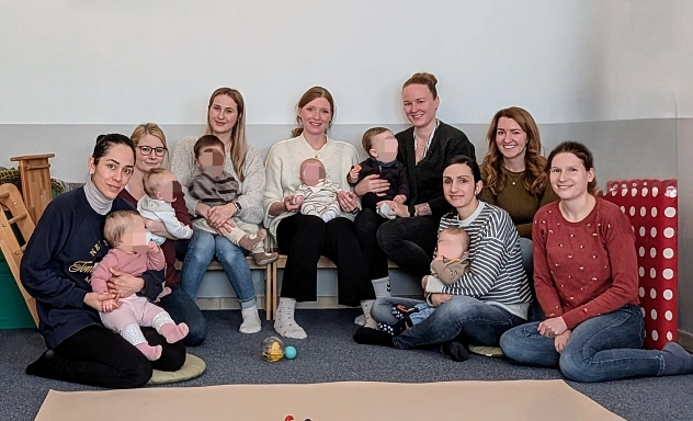 Eine Gruppe von acht Frauen sitzt gemeinsam mit ihren Babys in einem freundlichen Raum mit Teppichboden. Einige Mütter halten ihre Kinder auf dem Schoß, während andere auf einer Bank oder dem Boden sitzen. Die Babys tragen bequeme Kleidung und einige schauen neugierig umher. Im Hintergrund sind eine Kletterstruktur aus Holz und ein gepolstertes Element mit roten und weißen Punkten zu sehen. Die Atmosphäre wirkt warm und einladend.