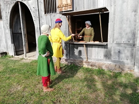 Drei mittelalterlich gekleidete Personen stehen vor dem Mittelalterhaus Nienover. Eine Person reicht an einem geöffneten Fenster etwas an eine weitere Person weiter.&nbsp;&copy;&nbsp;Das Rudel e. V.