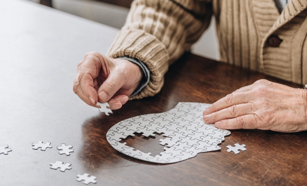 Zu sehen sind die Hände einer alten Person, die ein Puzzle in Form eines Kopfes zu vervollständigen versucht.