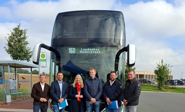 Doppeldeckerbus Landesbuslinie 160 Sechs Personen stehen vor einem großen Doppeldeckerbus mit der Aufschrift 160 Landesbus Duderstadt. Sie posieren für ein Gruppenfoto an einer Bushaltestelle bei sonnigem Wetter. Im Hintergrund sind ein Fahrradständer, Bäume und Gebäude zu sehen.