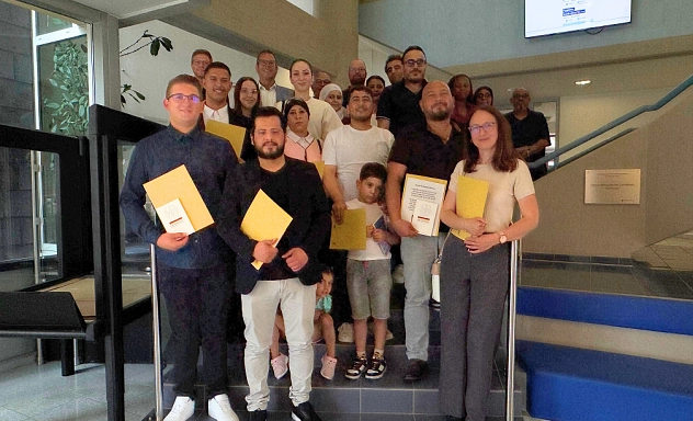 Einbürgerung Juni 2025 Eine Gruppe von Menschen steht auf einer Treppe in einem öffentlichen Gebäude und blickt in die Kamera. Einige halten gelbe Mappen und Urkunden in der Hand. Unter ihnen sind auch Kinder. Im Hintergrund steht Erster Kreisrat Jörg Richert. Über der Gruppe hängt ein Bildschirm mit der Aufschrift ‚Herzlich Willkommen‘. Das Foto wurde nach der feierlichen Übergabe der Einbürgerungsurkunden aufgenommen.“