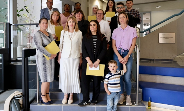 Einbürgerung Juni 2026.jpg Gruppenfoto auf einer Treppe im Foyer eines öffentlichen Gebäudes. Mehrere Erwachsene unterschiedlicher Herkunft stehen mit Urkundenmappen in den Händen für ein Erinnerungsfoto zusammen. Im Vordergrund steht ein kleines Kind. Die Gruppe blickt in die Kamera.