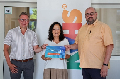 Drei Personen – zwei Männer und eine Frau – stehen vor einem Roll-up der Kreisvolkshochschule Northeim und halten gemeinsam ein Schild mit der Aufschrift ‚Wir sind Erasmus+ akkreditiert‘ in die Kamera. Das Foto entstand anlässlich der Erasmus+-Akkreditierung der KVHS.&nbsp;&copy;&nbsp;Landkreis Northeim