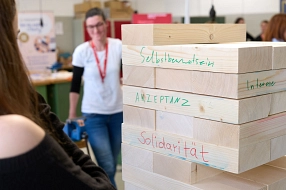 Nahaufnahme eines großen Holzklotz-Spiels, auf dessen Bausteinen Begriffe wie „Selbstbewusstsein“, „Akzeptanz“ und „Solidarität“ geschrieben stehen. Im unscharfen Hintergrund stehen zwei Personen an dem Spiel und beobachten die Aktion.&nbsp;&copy;&nbsp;EU-KOM