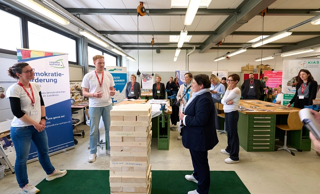 Europa in meiner Region Einbeck 1 Eine Gruppe von Menschen steht in einer Werkstatt an einem Informationsstand zur Demokratieförderung. Im Mittelpunkt erklärt ein Mann vor einem großen Holzklotz-Spiel etwas, während die Umstehenden aufmerksam zuhören. Auf mehreren Roll-ups sind Angebote des Landkreises Northeim und weiterer Einrichtungen zu sehen.