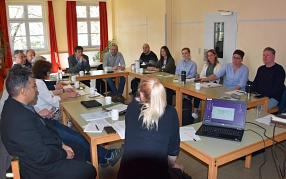 Fachleute am Runden Tisch Radverkehr In einem hellen Besprechungsraum mit großen Fenstern und roten Vorhängen sitzen rund 14 Personen an einem rechteckig angeordneten Tisch. Die Teilnehmenden wirken konzentriert und hören einer Person mit blonden Haaren im Vordergrund zu, die mit dem Rücken zur Kamera sitzt. Auf den Tischen liegen Dokumente, Notizblöcke, Stifte und Kaffeetassen, ein aufgeklappter Laptop zeigt eine geöffnete E-Mail-Anwendung. Die Atmosphäre deutet auf eine konstruktive Arbeitsbesprechung oder einen Workshop hin. © Landkreis Northeim