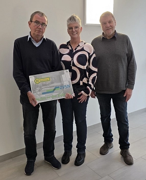 Drei Personen stehen nebeneinander in einem hellen Raum und blicken in die Kamera. Die Person links hält ein Schild mit dem Logo des Zweckverbandes Verkehrsverbund Süd-Niedersachsen (ZVSN) und der Aufschrift „Nächster Halt“. Die mittlere Person trägt eine gemusterte Bluse, die beiden Männer links und rechts sind in dunkler Freizeitkleidung gekleidet. Alle drei lächeln.&nbsp;&copy;&nbsp;ZVSN