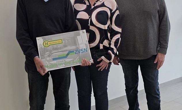 Fahrgastbeirat ZVSN Drei Personen stehen nebeneinander in einem hellen Raum und blicken in die Kamera. Die Person links hält ein Schild mit dem Logo des Zweckverbandes Verkehrsverbund Süd-Niedersachsen (ZVSN) und der Aufschrift „Nächster Halt“. Die mittlere Person trägt eine gemusterte Bluse, die beiden Männer links und rechts sind in dunkler Freizeitkleidung gekleidet. Alle drei lächeln.