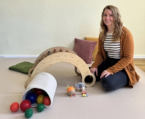 Familienbegleiterin Anne Lixfeld Familienbegleiterin Anne Lixfeld in einem gemütlich gestalteten Spielbereich. Vor ihr liegen verschiedene Spielsachen, darunter ein Holz-Kletterbogen, bunte Igelbälle und motorikförderndes Spielzeug für Kleinkinder. © Landkreis Northeim