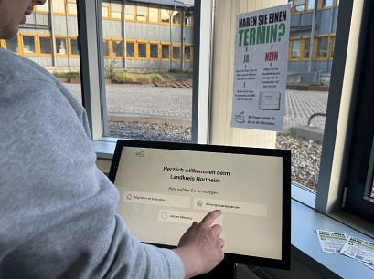 Zu sehen ist ein Front Desk Terminal zum digitalen Einchecken ins Kreishaus. Es steht in einem Raum mit Fenstern, links im Bild ist der Arm einer Person, die das Touch Screen berührt. Im Hintergrund hängt ein Plakat mit einer Anleitung wie man vorgeht, wenn man bereits einen Termin hat, bzw. wie man vorgeht, wenn man erst einen Termin vereinbaren möchte.&nbsp;&copy;&nbsp;Landkreis Northeim