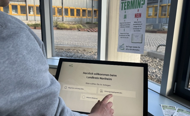 Front Desk Terminal Zu sehen ist ein Front Desk Terminal zum digitalen Einchecken ins Kreishaus. Es steht in einem Raum mit Fenstern, links im Bild ist der Arm einer Person, die das Touch Screen berührt. Im Hintergrund hängt ein Plakat mit einer Anleitung wie man vorgeht, wenn man bereits einen Termin hat, bzw. wie man vorgeht, wenn man erst einen Termin vereinbaren möchte.