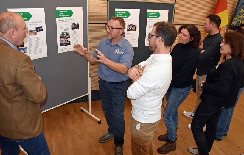 Zu sehen sind vier Personen die vor einer Pinnwand mit Informationen zum Thema Grüne Hausnummer stehen. Sie betrachten diese und sprechen über diese Informationen.