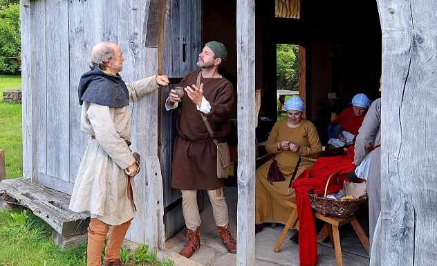 Gruppe Vuozvolc Vor dem Eingang eines mittelalterlichen Holzhauses unterhalten sich zwei Männer in historischer Kleidung. Im Inneren sitzen mehrere weitere Personen in Gewandung und arbeiten an Textilien. Die Szene vermittelt einen Einblick in den Alltag und das gemeinschaftliche Leben im Mittelalter.