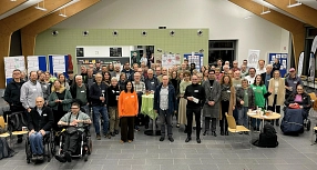 Die Teilnehmenden des Barcamps stehen und sitzen in einem großen, hell beleuchteten Raum der BBS Einbeck für ein Gruppenfoto zusammen. Im Hintergrund sind Stellwände mit Plakaten und Informationsmaterialien zu sehen. In der Mitte des Raumes steht ein runder Tisch mit grüner Tischdecke. Die Teilnehmenden blicken in die Kamera, viele lächeln.&nbsp;&copy;&nbsp;Landkreis Northeim