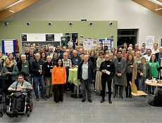 Gruppenbild Barcamp Demokratie 2025 Die Teilnehmenden des Barcamps stehen und sitzen in einem großen, hell beleuchteten Raum der BBS Einbeck für ein Gruppenfoto zusammen. Im Hintergrund sind Stellwände mit Plakaten und Informationsmaterialien zu sehen. In der Mitte des Raumes steht ein runder Tisch mit grüner Tischdecke. Die Teilnehmenden blicken in die Kamera, viele lächeln.