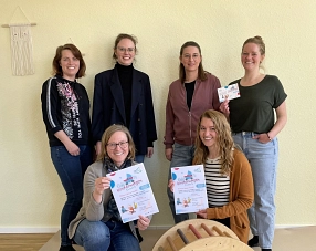 Eine Gruppe von sechs Frauen steht und kniet in einem freundlich gestalteten Raum vor einer beigefarbenen Wand mit Makramee-Dekoration. Zwei von ihnen halten Flyer in den Händen, die auf das "Café Kinderwagen Extra" hinweisen. Sie lächeln in die Kamera. Im Vordergrund ist ein Holzspielgerät mit bunten Stäben zu sehen.&nbsp;&copy;&nbsp;Landkreis Northeim