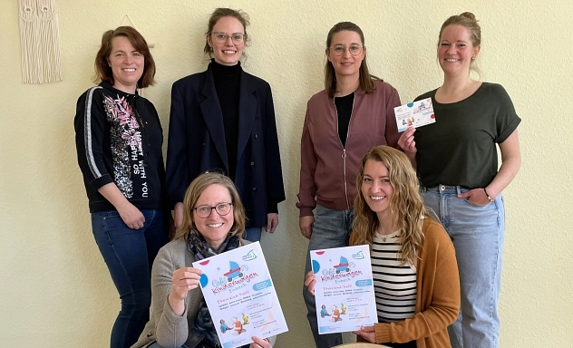 Eine Gruppe von sechs Frauen steht und kniet in einem freundlich gestalteten Raum vor einer beigefarbenen Wand mit Makramee-Dekoration. Zwei von ihnen halten Flyer in den Händen, die auf das "Café Kinderwagen Extra" hinweisen. Sie lächeln in die Kamera. Im Vordergrund ist ein Holzspielgerät mit bunten Stäben zu sehen.