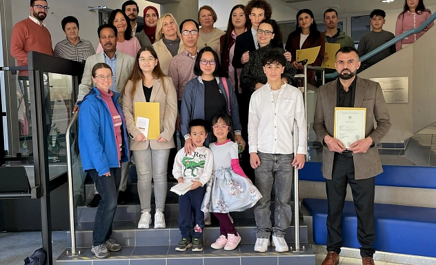 Gruppenbild Einbürgerung November 2025 Eine Gruppe neu eingebürgerter Menschen steht auf der Treppe im Eingangsbereich des Kreishauses Northeim. Vorne sind mehrere Kinder und Erwachsene zu sehen, einige halten ihre Einbürgerungsunterlagen in den Händen. Die Gruppe lächelt in die Kamera. Im Hintergrund erkennt man das Treppengeländer, Teile des Foyers und eine Wandtafel des Landkreises Northeim.