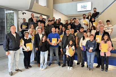 Eine große Gruppe von Menschen, bestehend aus Erwachsenen und Kindern, steht in einem hellen, modernen Treppenhaus. Viele der Anwesenden halten gelbe Mappen oder Unterlagen in der Hand. Die Stimmung wirkt freundlich und feierlich. Im Hintergrund ist auf einem Monitor ein Willkommensgruß zu sehen. Einige Personen stehen auf den Treppenstufen, andere auf dem Boden davor, sodass alle gut sichtbar sind. Die Gruppe setzt sich aus Menschen verschiedener Altersgruppen und Herkunft zusammen.&nbsp;&copy;&nbsp;Landkreis Northeim