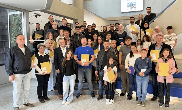 Gruppenbild Einbürgerungsfeier April 2025 Eine große Gruppe von Menschen, bestehend aus Erwachsenen und Kindern, steht in einem hellen, modernen Treppenhaus. Viele der Anwesenden halten gelbe Mappen oder Unterlagen in der Hand. Die Stimmung wirkt freundlich und feierlich. Im Hintergrund ist auf einem Monitor ein Willkommensgruß zu sehen. Einige Personen stehen auf den Treppenstufen, andere auf dem Boden davor, sodass alle gut sichtbar sind. Die Gruppe setzt sich aus Menschen verschiedener Altersgruppen und Herkunft zusammen.