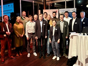 Das Bild zeigt eine Gruppe von Personen, die in einem Konferenzraum vor mehreren Bannern posieren. Die Gruppe besteht aus Männern und Frauen unterschiedlichen Alters, die überwiegend geschäftlich gekleidet sind. Im Hintergrund sind die Logos der Wirtschaftsförderung und andere Veranstaltungsinformationen sichtbar. Einige Personen tragen Namensschilder. Die Atmosphäre wirkt freundlich und professionell. Rechts im Bild ist ein Stehtisch mit Unterlagen und Technik zu sehen.&nbsp;&copy;&nbsp;Landkreis Northeim
