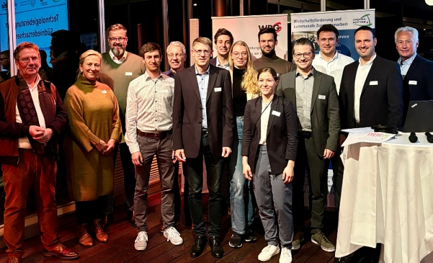 Gruppenfoto Informationsveranstaltung Technologie Das Bild zeigt eine Gruppe von Personen, die in einem Konferenzraum vor mehreren Bannern posieren. Die Gruppe besteht aus Männern und Frauen unterschiedlichen Alters, die überwiegend geschäftlich gekleidet sind. Im Hintergrund sind die Logos der Wirtschaftsförderung und andere Veranstaltungsinformationen sichtbar. Einige Personen tragen Namensschilder. Die Atmosphäre wirkt freundlich und professionell. Rechts im Bild ist ein Stehtisch mit Unterlagen und Technik zu sehen.
