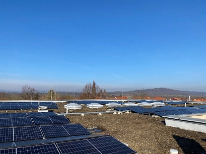 Mehrere Photovoltaikmodule auf dem Flachdach eines Gebäudes. Zwischen den Solarmodulen sind Dachfenster und technische Aufbauten zu sehen. Im Hintergrund erkennt man Dächer eines Ortes und den Turm einer Kirche vor einer hügeligen Landschaft unter blauem Himmel.&nbsp;&copy;&nbsp;Landkreis Northeim