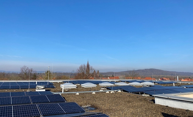 Inbetriebnahme PV-Anlage Schule Nörten-Hardenberg Mehrere Photovoltaikmodule auf dem Flachdach eines Gebäudes. Zwischen den Solarmodulen sind Dachfenster und technische Aufbauten zu sehen. Im Hintergrund erkennt man Dächer eines Ortes und den Turm einer Kirche vor einer hügeligen Landschaft unter blauem Himmel.